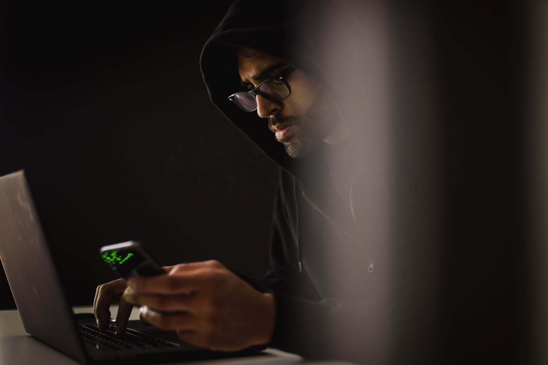crop ethnic hacker with smartphone typing on laptop in darkness
